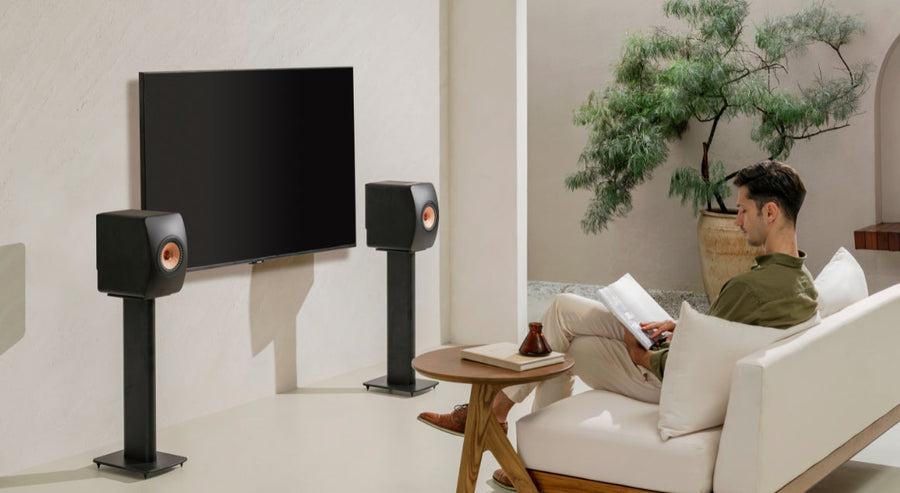 Homme détendu sur un canapé blanc dans un salon moderne, lisant un magazine, entouré de deux enceintes stéréo KEF sur pieds de chaque côté d’un téléviseur à écran plat, illustrant une installation audio haut de game.