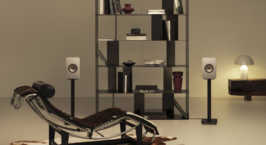 Salon minimaliste moderne avec deux enceintes bibliothèques blanches sur pieds noirs, une étagère ouverte noire avec livres et objets décoratifs, un fauteuil lounge design et une lampe de table à lumière douce.