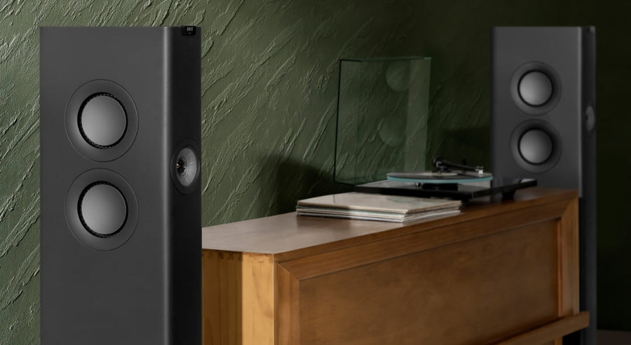 Deux enceintes colonnes modernes placées de chaque côté d’un meuble en bois avec une platine vinyle et des disques, devant un mur vert foncé à la texture marquée.