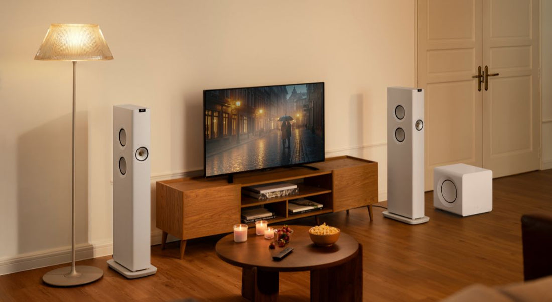 A home theater setup in a living room featuring two tall white floor-standing speakers, a white subwoofer, a TV on a wooden console, and a floor lamp.