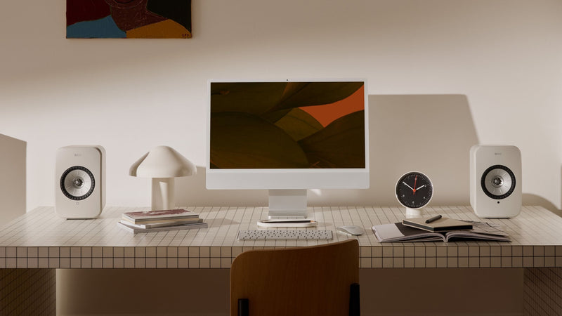 A desk featuring a computer, KEF LSX II LT Bookshelf speakers, and a stack of books, creating a workspace environment