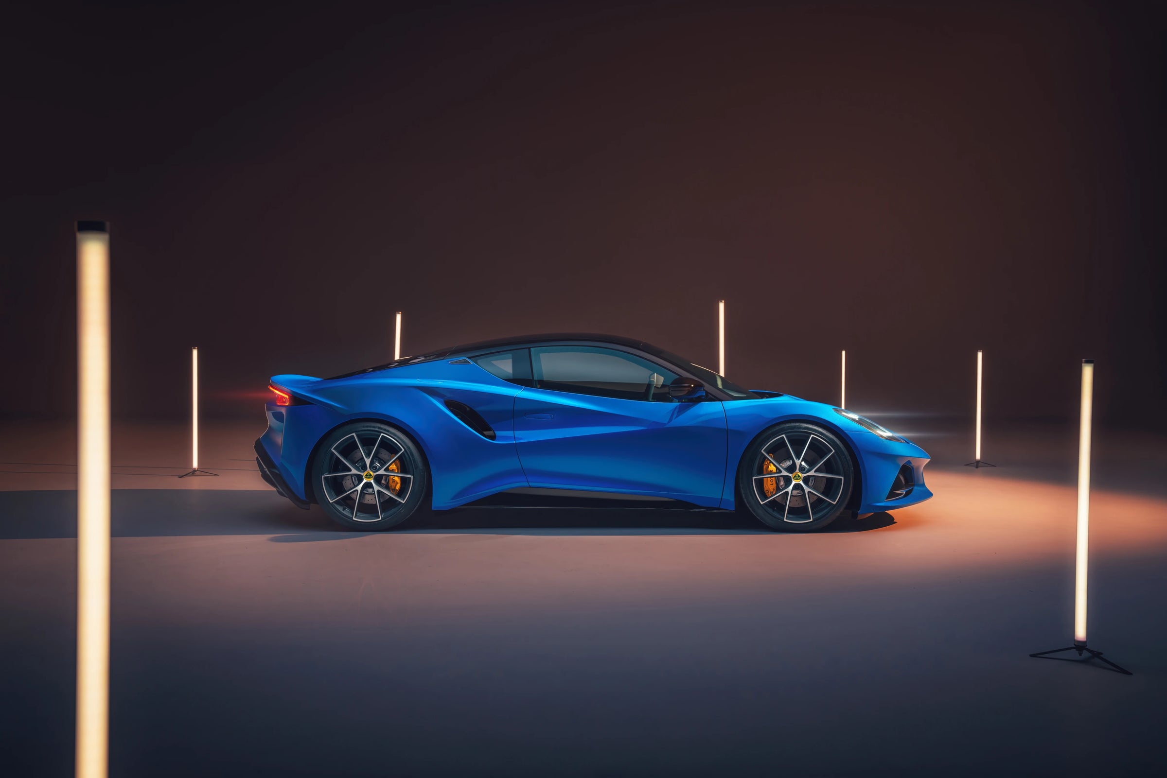 A bright blue Lotus Emira car shown in side profile, photographed in a dark studio setting with vertical light tubes casting warm light on the floor.